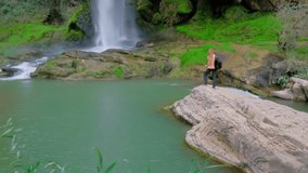 Woman is standing and raising her hands up. Cheering. Girl is hiking in nature. Green forest landscape with a waterfall. Horizontal video in a slow motion.