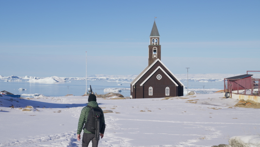 A wide-shot of a man hiking on a small path on the snow at the back of Zion Church in Ilulissat