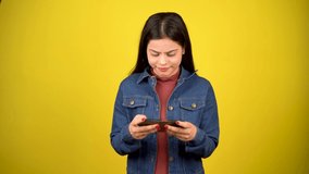 Portrait of happy modern girl using mobile phone, young woman playing video game on smartphone, standing isolated over yellow background