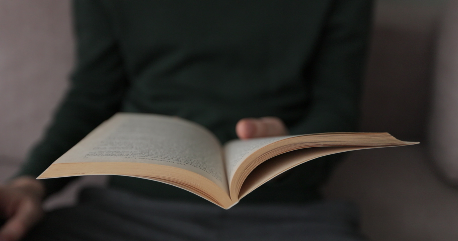 Man is lifestyle sitting on couch in cozy room reading favorite book. Man opens the book and turns the pages of the book.