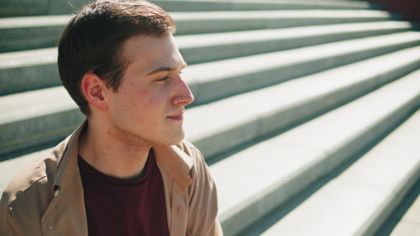 Young man portrait. Happy guy smiling close-up. Male cheerful student model posing, sitting on stairs outdoor, looking at camera.