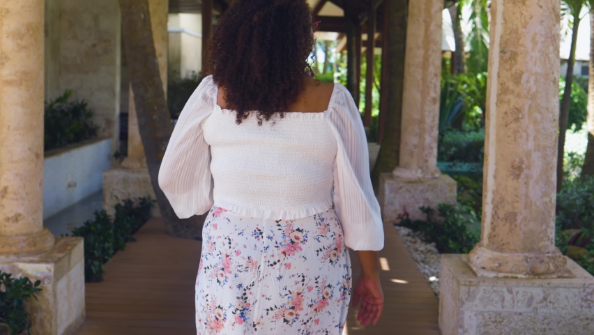 Black woman is walking around in pretty white floral dress during bright sunny day. she has curly hair and bright smile which shows how happy she is 