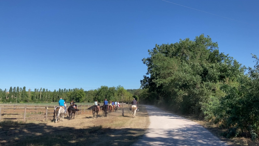 Rural scenery at Abbadia di Fiastra with a vision from behind of people horse riding on the large meadows of the abbey, with spinning tails, dust, a path, green trees, a contrail, and cars in the back