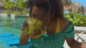 black woman sitting by the poolside drinking coconut water and enjoying the sun happily while on vacation. wearing a green summer dress and has curly brown hair its relaxing in the resort 