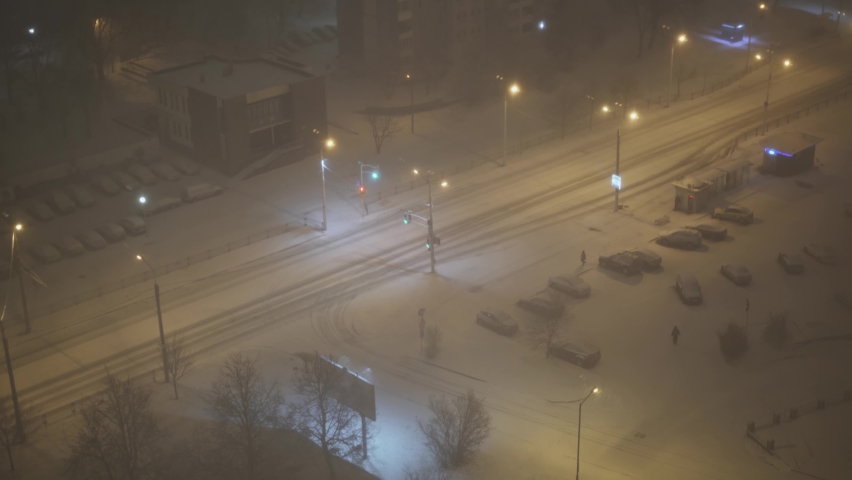 Deserted city street in snow storm at night, top view. Snow-covered car drives along road with street lighting. Few people on street, person in warm clothes walks through snow-covered parking lot.