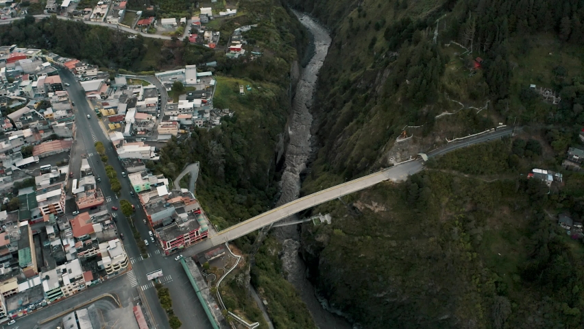 Aerial drone backward moving shot over a bridge on Pastaza river beside colorful City of Banos in Ecuador Settled on Basaltic Rock surrounded by mountains at daytime.