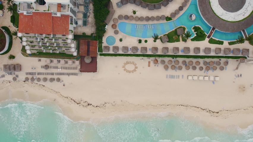 Aerial top down view of relaxing beach, crystal blue pools, and gentle waves at beautiful luxury resort Paradisus Cancun in Mexico with tiki huts and bird flying over beach.