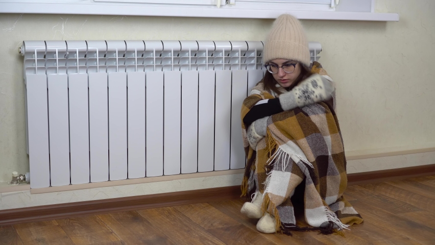 A young woman is sitting on the floor by the radiator covered with a blanket. The girl has not paid her heating bills and is sitting in warm clothes at home. | Shutterstock HD Video #1097162523