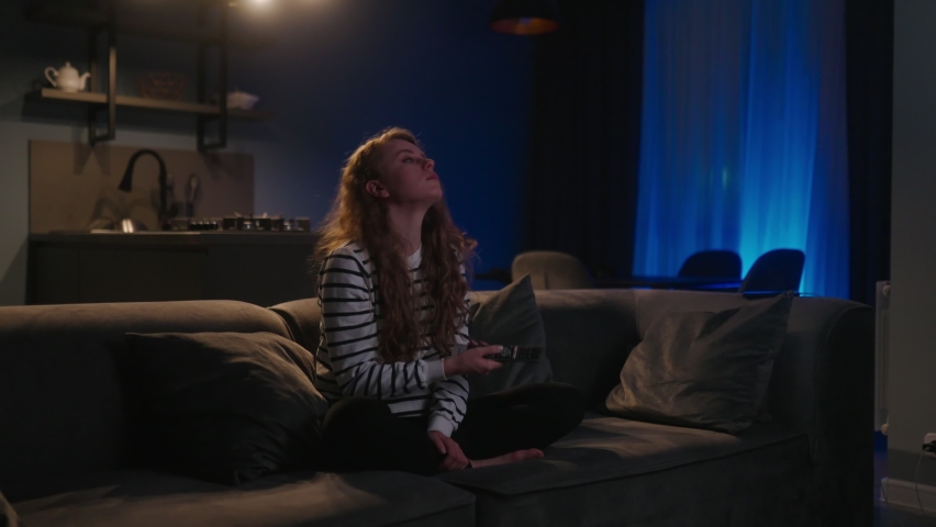 Cute blonde curly woman switching on television, navigating in smart tv applications while choosing channels to watch for entertain herself while sitting on sofa in living room at home. Cinematic shot | Shutterstock HD Video #1097164805