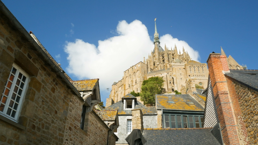 The gothic abbey of Mont Saint Michel viewed between two traditional and medieval buildings, Normandy, France. Blue sky on the background with white clouds.