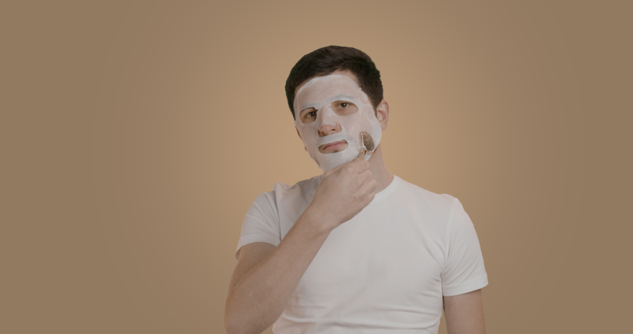 Portrait of a man doing a jade roller massage on his face wearing a cosmetic facial mask. A young man is doing a facial skin care treatment. Cosmetic procedures. | Shutterstock HD Video #1097166917