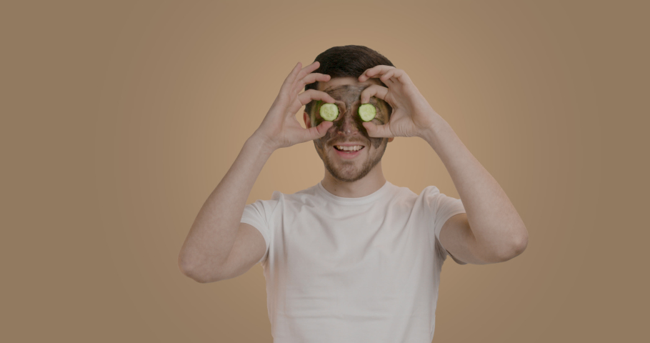 A man who has applied a black mask to his face and is waiting for it to take effect. The young man holds two cucumber rolls to his eyes and is amused and smiles widely. Facial procedures for men | Shutterstock HD Video #1097166919