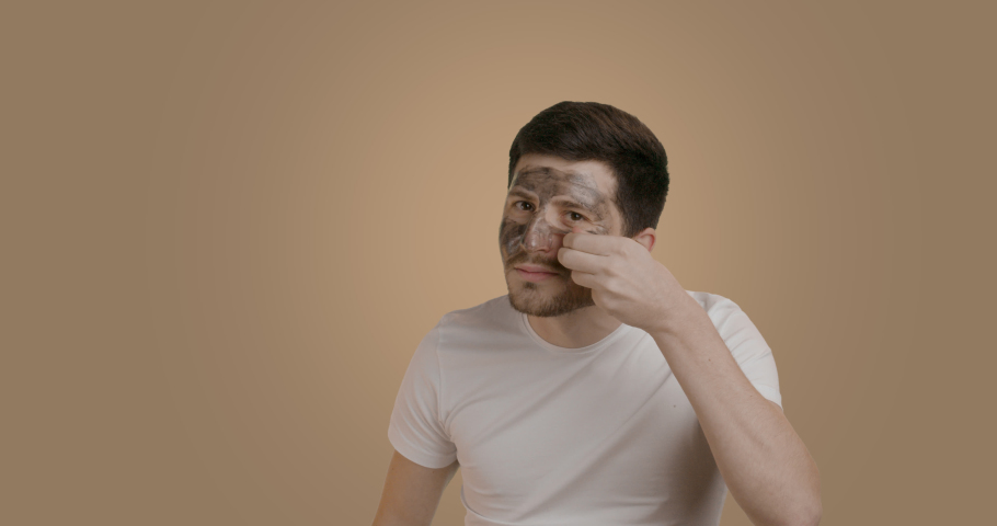 Smiling Caucasian man with a black mask on his face for black pores. The man takes off the face mask and shows what result he got - the concept of beauty and skin care. | Shutterstock HD Video #1097166921