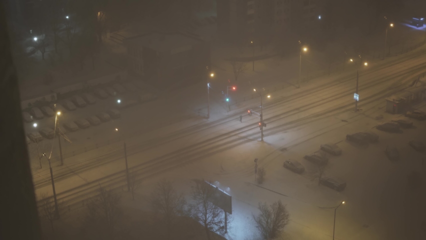 Heavy snowfall on city street at night, top view. Cars drive on snow-covered road under light of lanterns. Man in a warm jacket crosses the road. Blizzard in winter.