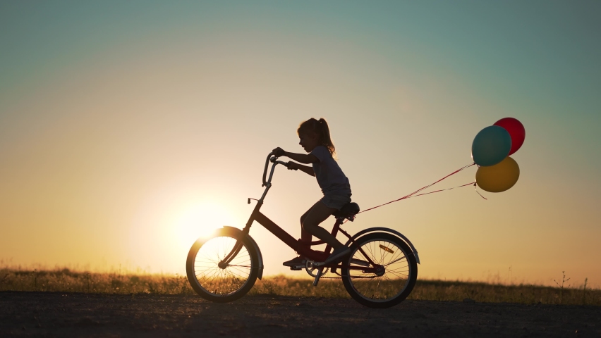 Dream kid. Silhouette of kid on bike in park. Girl rides in park on green grass. Child games in nature.Traveling with balloons on bike.Active child freedom in summer.Girl learns to ride bike in nature
