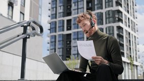 Modern hipster millennial urban man sit on stairs of epic office building, work on laptop remotely from office wearing headset reading document doing job. Freelance worker, project on the go outdoor. 