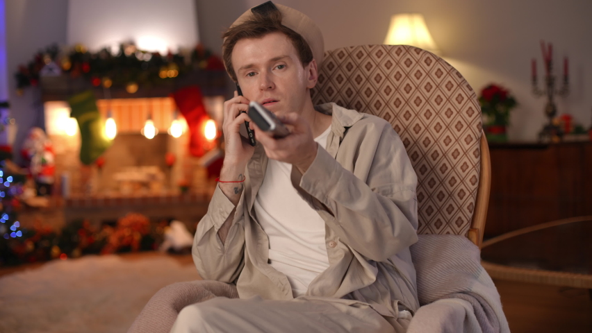 Absorbed Caucasian man talking on phone looking at camera switching TV channels with remote control. Portrait of concentrated LGBT guy chatting on Christmas eve at home sitting in living room | Shutterstock HD Video #1097176157