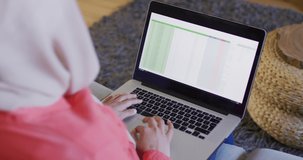 Over shoulder video of biracial woman in hijab at home working on laptop, spreadsheet on screen. Remote working, communication, inclusivity and domestic life.