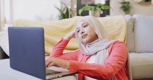 Video of concentrating biracial woman in hijab at home sitting on floor working on laptop. Remote working, communication, inclusivity and domestic life.