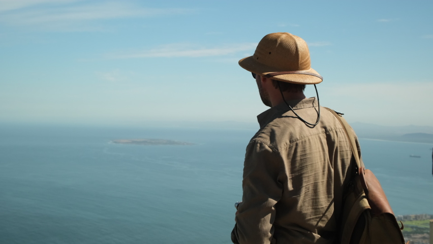 a male traveler in a sun hat points a finger at a famous island in the ocean. hiker man stands on a high mountain against the backdrop of the atlantic ocean. Robben Island, Cape Town, South Africa.