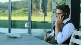 Inviting friends for dinner. Handsome young man in casual wear drinking wine and talking on the phone while sitting in a restaurant. Husband talking with her long-distance wife on a video chat