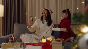Asian family merry xmas and happy new year holiday. Mom and daughter enjoy present gift christmas night at home. Mother and little girl sit on couch use smartphone video call surprise dad together.