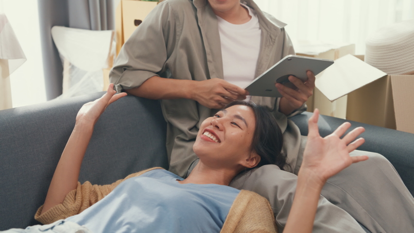 Young happy Asia couple sit on sofa couch use digital tablet online shopping furniture from decoration store in living room at home. Husband wife move new house, Apartment Rent And Ownership Concept. | Shutterstock HD Video #1097186855