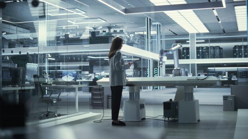 Industrial Robotics Engineer Comes to a Table With Robotic Arm in a High Tech Factory. Female Scientist Uses Tablet Computer to Manipulate and Program the Robot to Pick Up and Move a Metal Component.