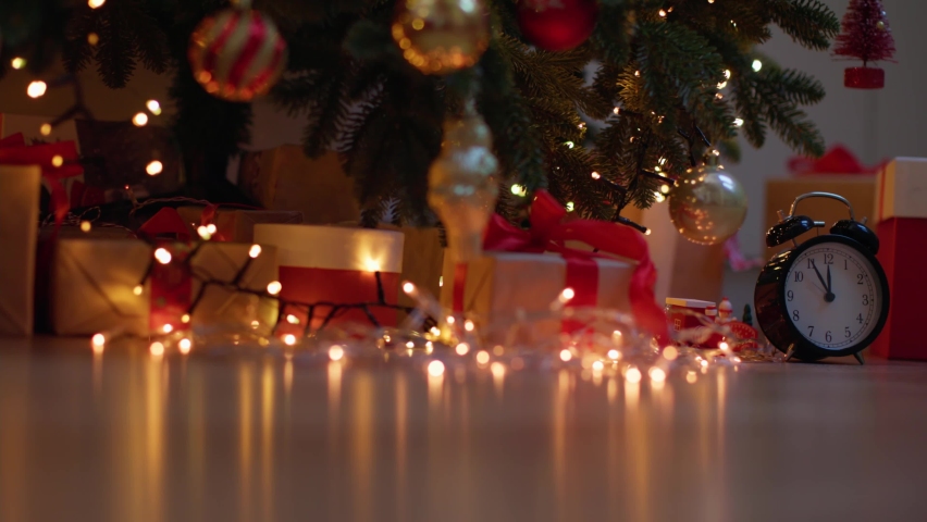 Many wrapped gift boxes alarm clock midnight time and christmas decorations under beautiful decorated christmas tree with golden bokeh lights in living room at night. craft brown paper gifts presents 
