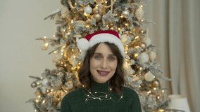  Young smiling woman wearing green sweater and Santa's cap calls her friends or parents via video conference call, saying hello and wishing Merry Christmas and Happy New Year. Xmas tree background