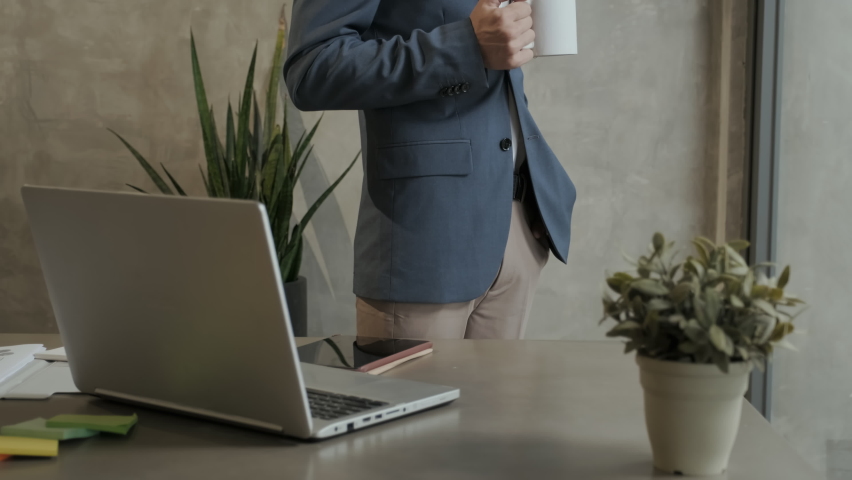 Tilt up of young successful Caucasian businessman in grey formalwear holding cup of coffee while standing by his workplace with laptop and looking at camera with smile | Shutterstock HD Video #1097194477