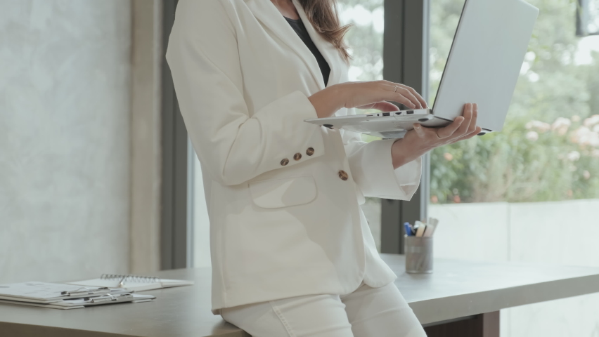 Tilt up of pretty young female employee in white suit standing by workplace in office, holding laptop, typing and looking at camera with smile | Shutterstock HD Video #1097196779