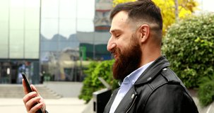 Positive Young Bearded Man Chatting while Standing on the Street. Video Chatting with Friends, Talking, Laughing via Modern Mobile Phone.