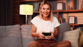 Portrait of excited middle aged blond woman gamer sitting on a couch and playing in video games on a console. Happy player female controlling joystick for online competition.