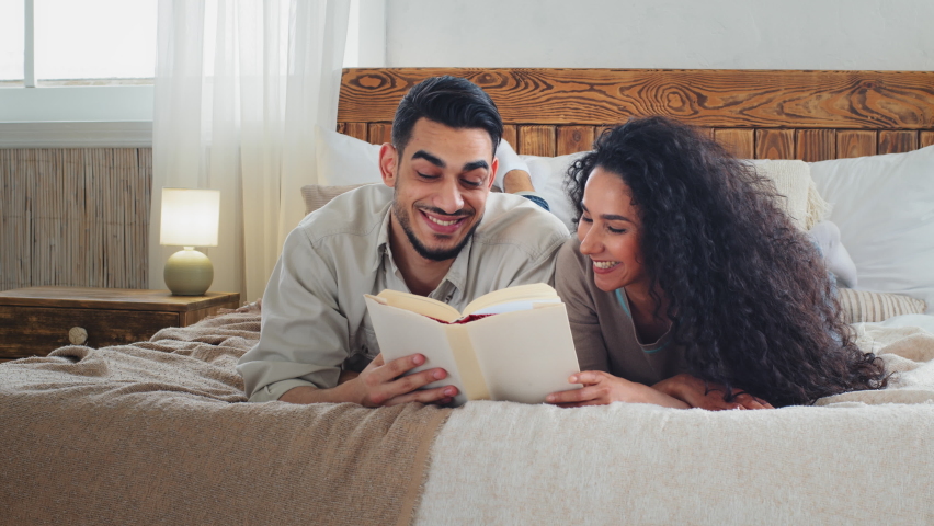 Ethnic multiracial multiethnic couple woman lady and bearded man lie together on bed read book. Married couple reading literature funny novel at home future parents choose name for child in textbook Royalty-Free Stock Footage #1097212643