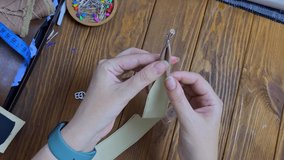 Women's fingers fold a ribbon of eco-leather in half and fasten it with clips. Top view of the sewing process on a wooden table. Hobbies and crafts.