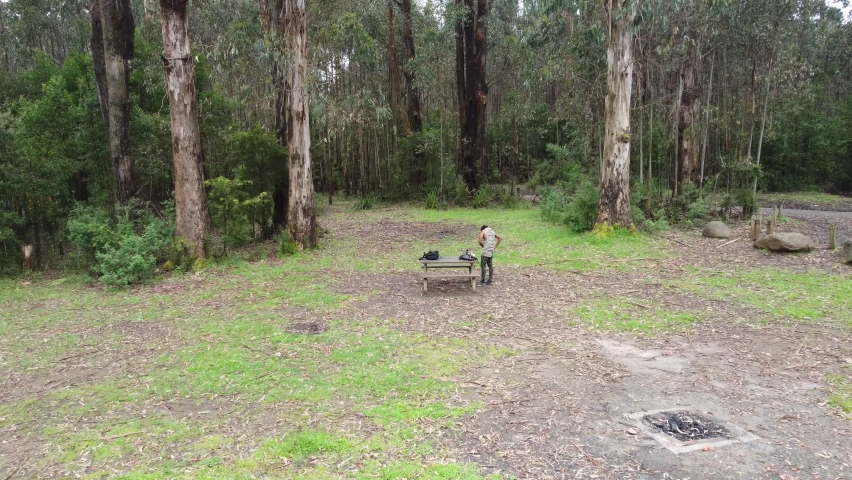 Aerial Drone Slow Cinematic shot of man packing camping and coffee items in a backpack on a picnic table in a park surrounded by tall trees