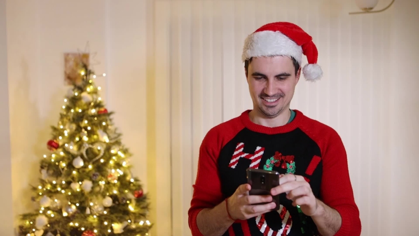 Christmassy dressed male with a Xmas jumper smiling browsing smartphone content in front of Christmas tree doing online shopping | Shutterstock HD Video #1097219759
