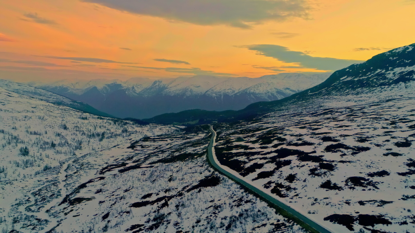 Aerial pullback over road cutting through snowy mountainous landscape - vibrant orange sunset sky in Norway