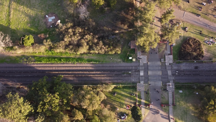 Aerial top down shot of train on rail driving next to Agronomia Park with resting people at sunset