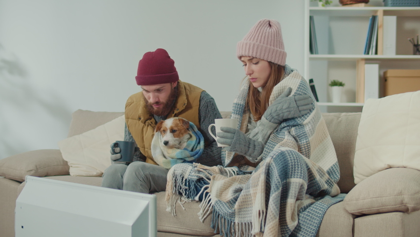 A Couple Try to Warm Up Their Dog in the Cold Room. A Couple with Their Dog in Cold Home Warmly Clothed Hugging Sitting on a Sofa at Home. Due Debt, Low Temperature Indoor Energy Crisis Concept. Royalty-Free Stock Footage #1097227639