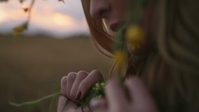 Beautiful woman with floral crown walks thoughtfully at wheat field, touches small wild flowers. Romantic enigmatic face, professional bright make up. Sunset out of city. High quality 4k