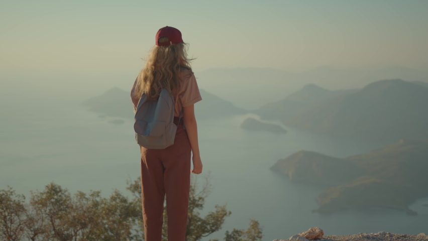 Child traveler joyfully raises up hands in mountains. School-age girl with backpack admires beautiful view of sea and mountains