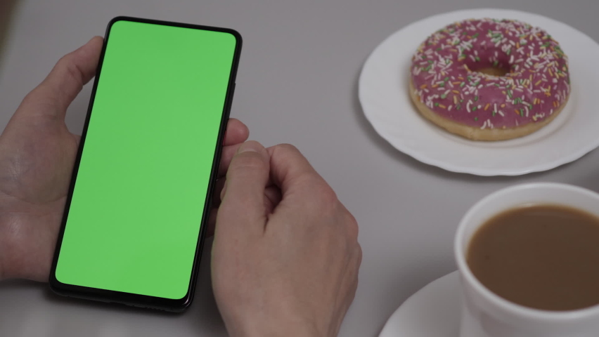 Woman Sitting at Table Coffee Donut Using Smartphone With Chroma Key Green Screen, Scrolling Through Social Network Media Online Shop Internet. Smartphone in Horizontal Mode with Green Screen Mock-up. | Shutterstock HD Video #1097232639