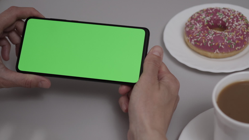 Woman Sitting at Table Coffee Donut Using Smartphone With Chroma Key Green Screen, Scrolling Through Social Network Media Online Shop Internet. Smartphone in Horizontal Mode with Green Screen Mock-up. | Shutterstock HD Video #1097232653