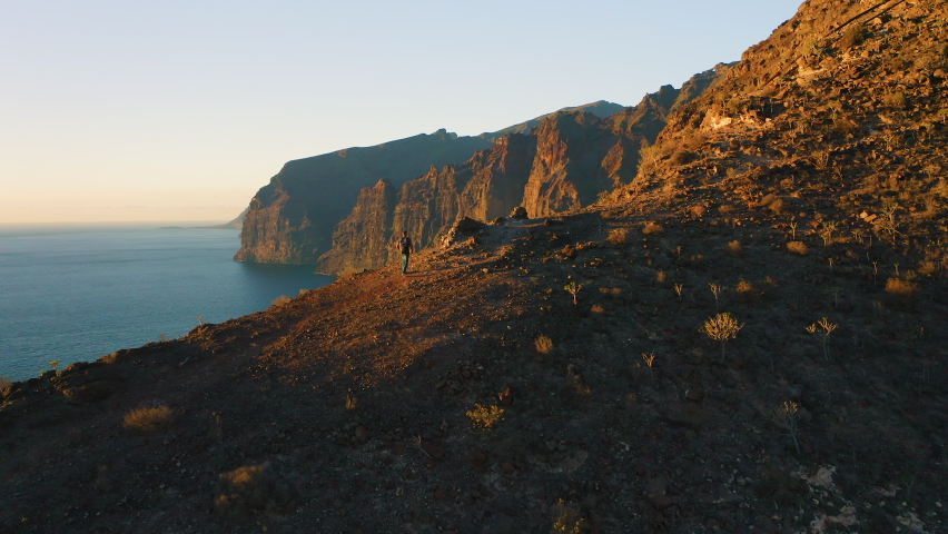 Man with backpack climbs the mountainside and contemplates ocean landscape from top of cliff at evening sky sunset background. Rear aerial view of tourist backpacker on rocky shoreline.