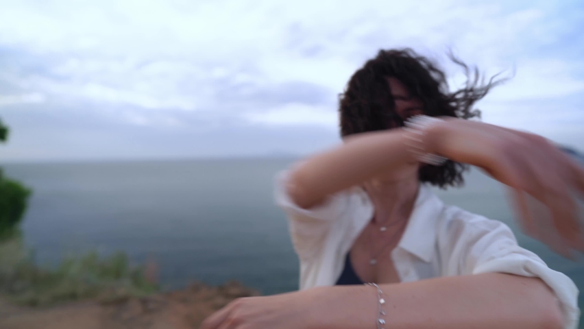 Lady with short dark wavy hair is dancing sensual performance standing on the cliff against sea. Caucasian woman is moving to rhythm with grace listening to music by wireless earphones. Freedom at art