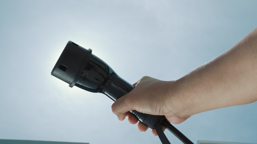 Closeup hand grasping an EV plug for electric vehicle with the midday sky in the background as progressive idea of alternative sustainable clean and green energy for environmental concern. | Shutterstock HD Video #1097240947