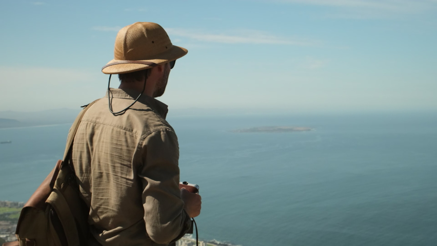 a male traveler in a sun hat points a finger at a famous island in the ocean. hiker man stands on a high mountain against the backdrop of the atlantic ocean. Robben Island, Cape Town, South Africa. Royalty-Free Stock Footage #1097244619