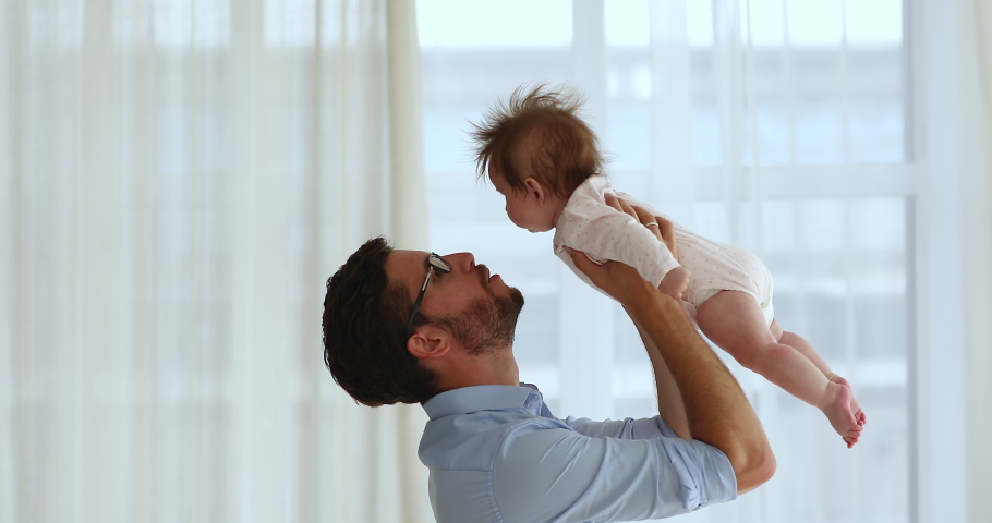 Cheerful happy playful new dad playing with baby at home window, cuddling, kissing, lifting little kid up in air, smiling, having fun, enjoying fatherhood. Side view. Childcare, family Royalty-Free Stock Footage #1097249787